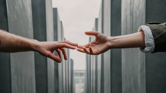 In der Mitte einer langen Reihe grauer Gebäude werden in einer unterstützenden Geste zwei Hände aufeinander zu gestreckt.  | © Toa Heftiba/unsplash