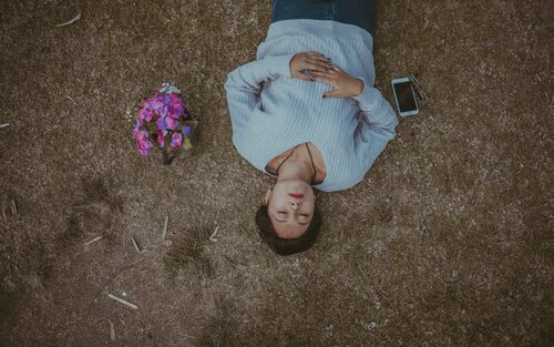 Frau liegt mit Händen auf dem Bauch und geschlossenen Augen am Boden. Daneben ein Blumenstraus. | © Christian Newman/unsplash