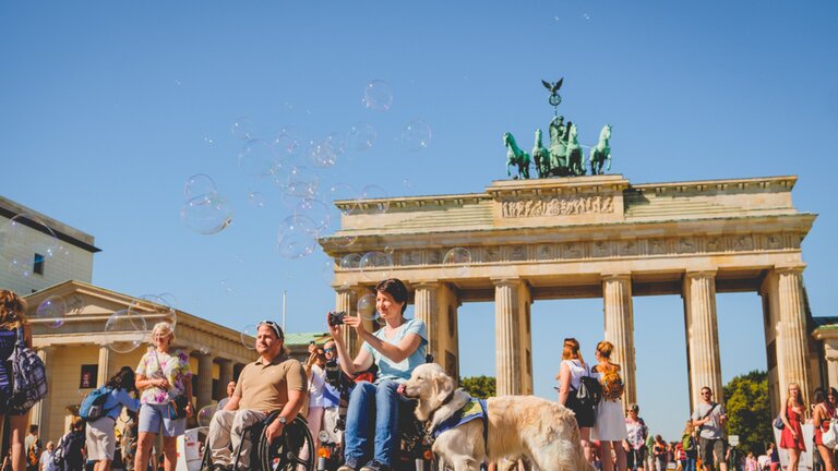 Es ist ein sonniger Tag und vor dem Brandenburger Tor in Berlin stehen viele verschiedene Leute. Sie schießen Fotos, pusten Seifenblasen in die Luft oder betrachten die Umgebung. In der Mitte des Bildes ist eine Frau im Rollstuhl, die eine Kamera in der Hand hält. Neben ihr steht ein Hund. | © Andi Weiland | Gesellschaftsbilder.de