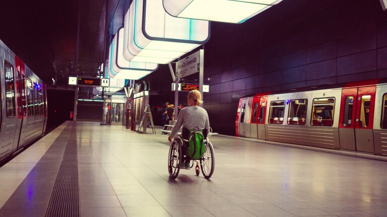 Eine Frau mit blonden Haaren befindet sich mit ihrem Rollstuhl in einer U-bahn Station bei der gerade zwei Bahnen auf den Gleisen stehen. | © Lukas Kapfer | www.th-10.de