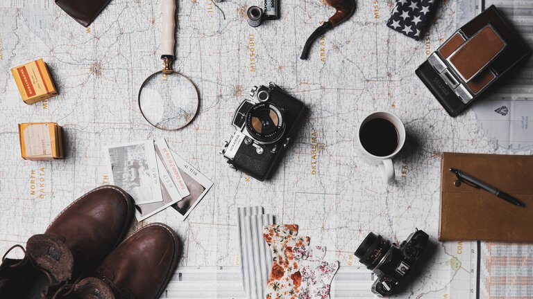 Weltkarte, auf der verschiedene Gegenstände wie Kameras, Schuhe, eine Lupe, eine Pfeife und vieles mehr liegen. | © Unsplash